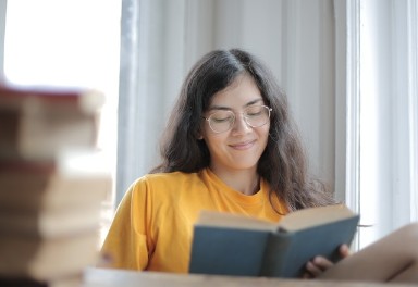 Een jonge vrouw met een geel t-shirt en een bril leest tevreden in een boek, terwijl ze aan een tafel zit waar een stapel met nog meer boeken ligt.